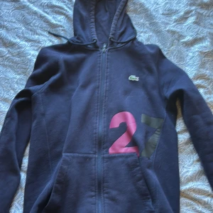  Lacoste Äkta tröja - Säljer en mörkblå hoodie från Lacoste Sport med dragkedja och nummerdetalj i rosa och svart på framsidan. Perfekt för en sportig look. Den har en justerbar huva och långa ärmar.