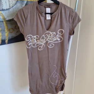 Brun kortärmad topp från Baby Phat - Snygg brun kortärmad topp från Baby Phat med glittrande text och kattmotiv i strass. Toppen har en v-ringad hals och är perfekt för en trendig look.