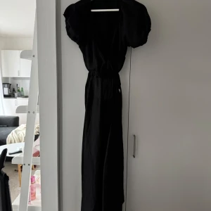 Svart klänning med puffärmar - Elegant svart klänning med puffiga kortärmar och omlott-design. Klänningen har en v-ringad hals och är tillverkad i ett lätt och luftigt material, perfekt för varmare dagar.