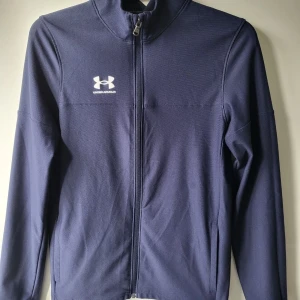  Under Armour tröja  - Säljer en mörkblå jacka från Under Armour med dragkedja och hög krage. Jackan har en stilren design med logotypen i vitt på bröstet. Perfekt för träning eller vardagsbruk.