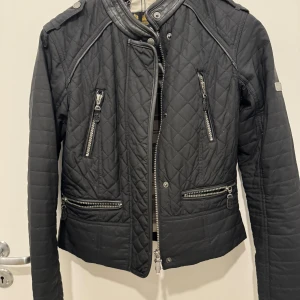 Svart quiltad jacka från Barbour - Säljer en snygg svart quiltad jacka från Barbour International. Jackan har en cool bikerstil med dragkedjor och en ståkrage med tryckknapp. Perfekt för en stilren look!