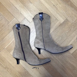 Beige mocka cowboy boots med klack - Snygga beige cowboy boots i mocka med en elegant klack. De har en dragkedja på sidan och svarta detaljer upptill. Perfekta för en stilren look.