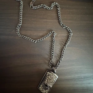 Silverfärgat halsband med dödskallehänge - Coolt silverfärgat halsband med ett rektangulärt hänge prytt med glittrande stenar och en dödskalle i mitten. Perfekt för att ge din outfit en edgy touch.