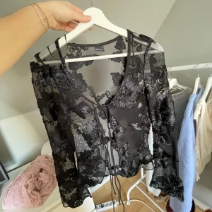 Svart blommig meshblus från Gina Tricot - Säljer en transparent svart meshblus från Gina Tricot med blommigt broderi och knyt framtill. Blusen har långa ärmar.