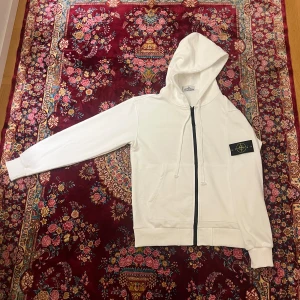 Vit hoodie från Stone Island - Vit hoodie från Stone Island med svart dragkedja och klassisk logotyp-patch på vänster ärm. Jackan har huva med dragsko och ribbade muddar. Perfekt för en stilren och avslappnad look, matchar sommar auran och sitter väldigt snyggt. Storleken är S men passa väldigt bra på M 