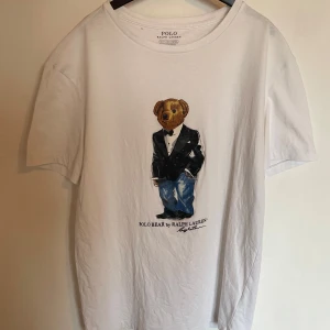 Vit t-shirt med björntryck från Polo Ralph Lauren - Vit t-shirt från Polo Ralph Lauren med ikoniskt björntryck på bröstet. Klassisk rund halsringning och normal passform. Perfekt för dig som gillar stilrena plagg. Finns ett litet hål se, (bild 2) därav priset. Passar dig som brukar ha XS. Skriv vid minsta fundering. Priset kan diskuteras vid snabbaffär!!