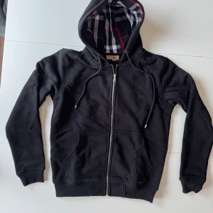 Svart hoodie från Burberry med dragkedja - Säljer en svart hoodie från Burberry med dragkedja och klassiskt rutigt foder i luvan. Tröjan har två fickor framtill, justerbar luva med snören och broderad logga på bröstet. Perfekt för en stilren och bekväm look. ((FRAKTAR SAMMA DAG)) 🎉 
