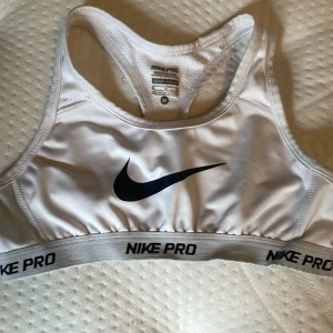Vit sport-bh från Nike Pro - Säljer en vit sport-bh från Nike Pro med svart logga framtill och resår med Nike Pro-tryck. Modellen har brottarrygg och är tillverkad i ett funktionsmaterial som andas. Perfekt för träning och sportaktiviteter.