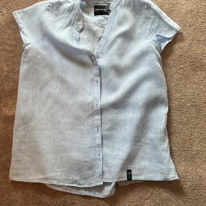 Ljusblå linneskjorta från Pelle P - Snygg ljusblå skjorta/blus från Pelle P i  linne. Skjortan har korta ärmar och knäppning framtill. Perfekt för varma dagar och enkel att styla med jeans eller shorts. Diskret broderad logga på ärmen.