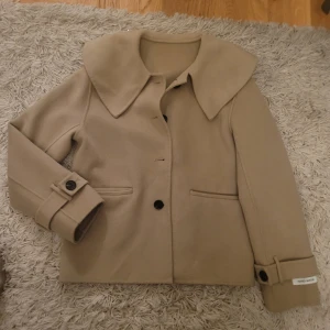 Beige kappa med stora krage - Säljer en supersnygg beige kappa med stora krage och knappar framtill. Jackan har långa ärmar med knappar vid ärmsluten. Pris kan diskuteras!