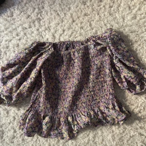 Blommig offshoulder blus från Vero Moda - Supersöt blommig offshoulder blus från Vero Moda i lila, gul och grön. Blusen har volangdetaljer nertill och vid ärmslut samt smockad midja. Perfekt för vår och sommar!