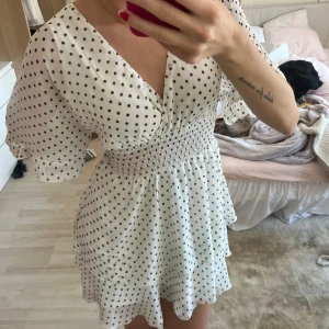 Vit kortklänning med svarta prickar - Supersöt vit kortklänning med svarta prickar och v-ringning. Klänningen har korta, vida ärmar och smock i midjan som ger en fin passform. Tillverkad i ett lätt och luftigt material, perfekt för varma dagar.