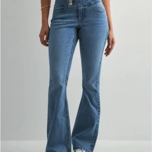 Blå bootcut jeans från Vero Moda - Säljer ett par klassiska blå bootcut jeans från Vero Moda i modellen VMSGI LR DB Button SK Flare Jeans. Jeansen har medium midja, två knappar framtill och är tillverkade i medium blue denim. Perfekta för dig som gillar en tidlös stil.