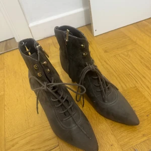 Högklackade ankle boots - Säljer ett par olivgröna boots med smal klack. Skorna har snörning framtill, dragkedja på sidan och guldfärgade detaljer vid snörhålen. Perfekta för dig som vill ha en stilren och edgy look.