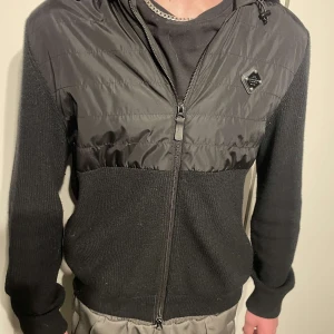 J.Lindeberg hybridjacka med huva - Säljer en svart j.Lindeberg jacka med stickade ärmar och nederdel samt glansig syntet på bröst och huva. Jackan har dragkedja framtill och en liten logodetalj på bröstet. Perfekt för dig som vill ha en sportig och stilren look. Storlek L men sitter som M