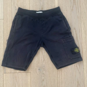 Svarta shorts från Stone Island Junior - Svarta shorts från Stone Island Junior med elastisk midja och sidoficka med ikonisk logotyp-patch. Klassisk passform och tillverkade i mjukt bomullsmaterial. Perfekta för en avslappnad stil.