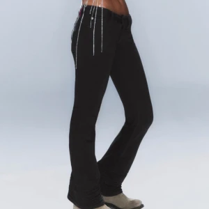 Svarta bootcut jeans med kedjedetaljer - Säljer ett par svarta bootcut jeans med låg midja och snygga kedjedetaljer som hänger från fickorna. Jeansen har klassisk femficksdesign och stängs med dragkedja och knapp. Perfekta för dig som vill ha en cool och trendig look.