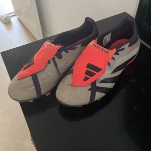 Adidas Predator fotbollsskor i grått och rött - Säljer ett par Adidas Predator fotbollsskor i grått med röda detaljer och svarta ränder. Skorna har täckt snörning och mönstrad sula för bra grepp på planen. Perfekta för dig som vill sticka ut på fotbollsplanen! Och priset kan diskuteras 
