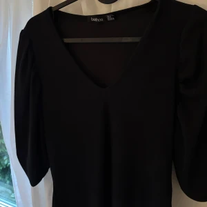 Svart topp med v-ringning från Boohoo - Säljer en svart topp från Boohoo med v-ringad hals och långa ärmar. Toppen har en enkel och stilren design som passar perfekt till olika tillfällen. Materialet känns mjukt och följsamt mot kroppen.