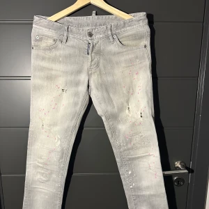 DSQUARDE2  ljusgrå jeans med färgstänk - Säljer ett par ljusgrå Replay jeans med unika rosa och vita färgstänk samt slitningar på benen. Byxorna har fem fickor, normal passform och klassisk dragkedja. Perfekta för dig som vill sticka ut med en kreativ och trendig look. Byxorna är i väldigt bra skick. Ni kan kolla på bild nummer två hur byxorna sitter på mig som är en och 165. Jag skulle starkt rekommendera dom här byxorna till dig som är runt 165-170.