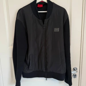 Svart jacka från Hugo  - Snygg svart jacka från Hugo  med ribbade muddar och krage. Jackan har dragkedja framtill, två fickor och en diskret logotyp på bröstet. Perfekt för en stilren och modern look.