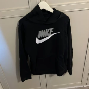 Svart hoodie från Nike - Svart hoodie från Nike med stor vit logga framtill. Klassisk modell med huva och magficka, perfekt för en sportig och avslappnad stil. Passar lika bra till jeans som till träningsbyxor.