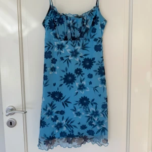 Blå blommig klänning från Antico Anna Concepts - Säljer en blå blommig klänning med smala axelband och lätt rynkad byst. Klänningen har en feminin passform och blommigt mönster i olika blå nyanser. Perfekt för varma dagar och somriga tillfällen.