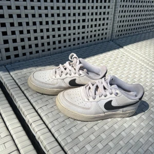 Vita Nike Air Force 1 sneakers med svart swoosh - Säljer ett par klassiska vita Nike Air Force 1 sneakers med svart swoosh och svart detalj på hälen. Skorna har snörning, rund tå och platt sula. Perfekta för dig som gillar streetstyle och vill ha en tidlös look. Även jätte enkla att tvätta. 
