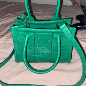 Grön handväska från Marc Jacobs - Säljer en snygg grön handväska från Marc Jacobs med präglad logga framtill. Väskan har både handtag och avtagbar axelrem samt dragkedja upptill. Perfekt för dig som vill ha en färgklick till outfiten!