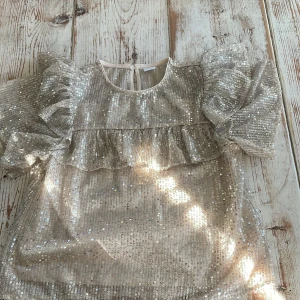 Glittrig topp med volang från Lindex - Säljer en glittrig topp från Lindex i ljusbeige med paljetter. Toppen har puffiga korta ärmar och volangdetalj framtill. Perfekt för dig som gillar att sticka ut med lite extra glitter!
