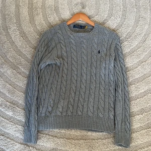 Grå stickad tröja från Polo Ralph Lauren - Säljer en klassisk grå kabelstickad tröja från Polo Ralph Lauren. Tröjan har rund halsringning, långa ärmar och den ikoniska broderade loggan på bröstet. Perfekt för lager-på-lager och enkel att matcha med det mesta.