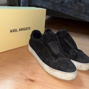Svarta sneakers från Axel Arigato i mocka - Säljer ett par svarta sneakers från Axel Arigato i mjuk mocka. Skorna har en stilren design utan snörning och med vit platt sula. Perfekta för dig som gillar minimalistisk stil och vill ha något som funkar till det mesta. 