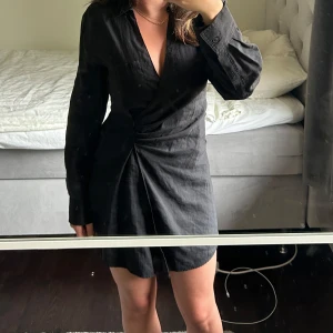 Svart omlottklänning med v-ringning - Snygg svart omlottklänning med v-ringning och långa ärmar. Klänningen har en kort längd och markerad midja som ger en smickrande siluett. Perfekt för dig som gillar stilrena och enkla plagg.