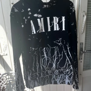 Svart hoodie från Amiri med vit mönster - Säljer en svart hoodie från Amiri med vit sprucken grafisk design och stor logga framtill. Tröjan har långa ärmar och en klassisk huva. Perfekt för dig som gillar streetwear och vill sticka ut med en unik look.  