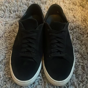 Svarta sneakers från Axel Arigato - Snygga svarta sneakers från Axel Arigato med vit platt sula och diskret logga på sidan. Skorna har snörning och är tillverkade i mocka för en stilren look. Perfekta för dig som gillar minimalistisk design. Skriv om ni undrar något✅