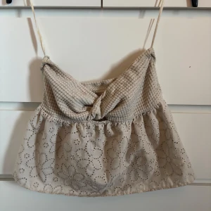 Beige croppad linnetopp med spetsdetaljer - Söt beige croppad topp. Överdelen är ribbad och har en knut framtill, medan nederdelen är dekorerad med broderade blommor i spets. Perfekt för varma dagar.