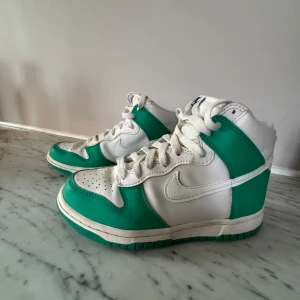 Nike Dunk High vit och grön - Knappt använda! Säljer ett par Nike Dunk High i vitt och grönt. Skorna har klassisk hög siluett, vit bas med gröna paneler och vit sula med grön undersida. Ikonisk Nike-logga på plösen och sidorna. Perfekta för dig som gillar streetwear och sneakers.