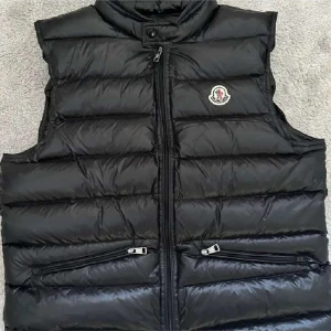 Svart dunväst från Moncler - Snygg svart dunväst från Moncler med hög krage och dragkedja framtill. Västen har två fickor med dragkedja och Moncler-logga på bröstet. Perfekt för lager-på-lager och stilren look.