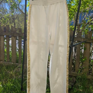 Vita byxor med guldiga paljettband - Säljer ett par vita byxor med bred resår i midjan och guldiga paljettband längs sidorna. Byxorna har en rak passform och är perfekta för dig som vill sticka ut lite extra.