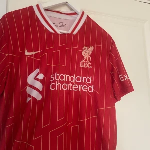 Liverpool FC hemmatröja 2023/24 - Röd Liverpool FC fotbollströja från Nike med vita och gula detaljer. Tröjan har vita ränder, klubbmärke och sponsortryck på bröstet samt Champions League-märke och Foundation-tryck på ärmen. Klassisk V-ringad krage med vita inslag.