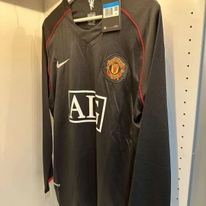 Manchester United långärmad svart fotbollströja - Säljer en svart långärmad Manchester United fotbollströja från Nike med AIG som sponsor på bröstet. Tröjan har röda detaljer längs axlarna och klubbmärket broderat på vänster bröst. Perfekt för fans som vill sticka ut på läktaren.