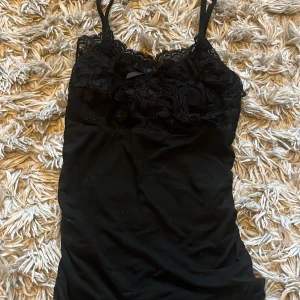 Svart spetslinne med rosett - Säljer ett svart linne med smala axelband och vacker spets upptill. Linnet har en liten rosett framtill och är figurnära i modellen. Perfekt att styla till olika outfits!