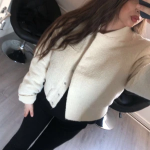 Beige croppad cardigan jacka - Säljer en beige croppad cardigan jacka med knappar framtill. Jackan har lång ärm, ribbade muddar och en mjuk, lite fluffig textur. Perfekt för att lyfta en enkel outfit med en stilren touch.