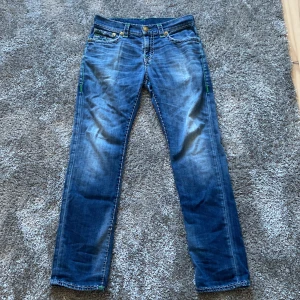True religion - Säljer ett par blå jeansbyxor från True Religion, modell GENO Relaxed Slim. Byxorna har gröna broderade detaljer på bakfickorna och klassisk femficksdesign. Snygg kontrastsöm och normal passform med slim siluett. Enda defekter är saknade knappar på bakfickorna.  Vid intresse tveka inte att fråga något. Pris går att diskutera vid snabb affär.