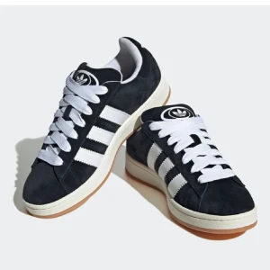 Adidas Campus svarta sneakers - Klassiska Adidas Campus sneakers i svart mocka med vita ränder och vit sula. Skorna har breda vita skosnören, gummisula och rund tå. Perfekta för en stilren och sportig look.