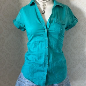 Turkos kortärmad skjorta med knappar - Snygg turkos skjorta med korta ärmar och knappar hela vägen framtill. Skjortan har en klassisk krage och en bröstficka med lock. Perfekt till jeans eller kjol för en fräsch look.