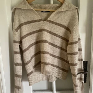 Beige och brun randig stickad tröja från Gina Tricot - Mysig stickad tröja från Gina Tricot i beige med bruna ränder. Tröjan har v-ringad krage och långa ärmar med slits och ribbade muddar.