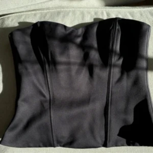 Svart korsettopp - Säljer en svart korsettopp med markerade sömmar framtill. Toppen har en figurnära passform och är axelbandslös, perfekt att styla till olika outfits.