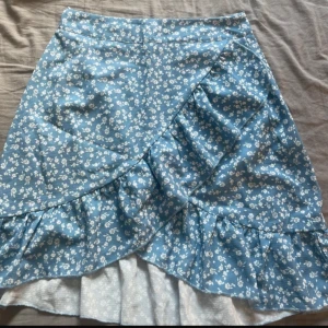 Blå blommig kjol med volang - Säljer en söt blå kjol med vitt blommönster och volangdetalj framtill. Kjolen har omlottkänsla och är perfekt för vår och sommar. Lätt och luftig modell som ger en romantisk vibe.