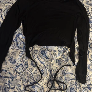 Svart croppad långärmad topp med knyt - Svart långärmad croppad topp med knytband i midjan. Enkel och stilren design som passar till många olika outfits. Mjukt och stretchigt material för en bekväm känsla.